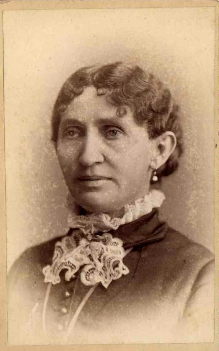A woman with light skin, curly hair pulled back, a lacy white collar, and white high-collared dark blouse
