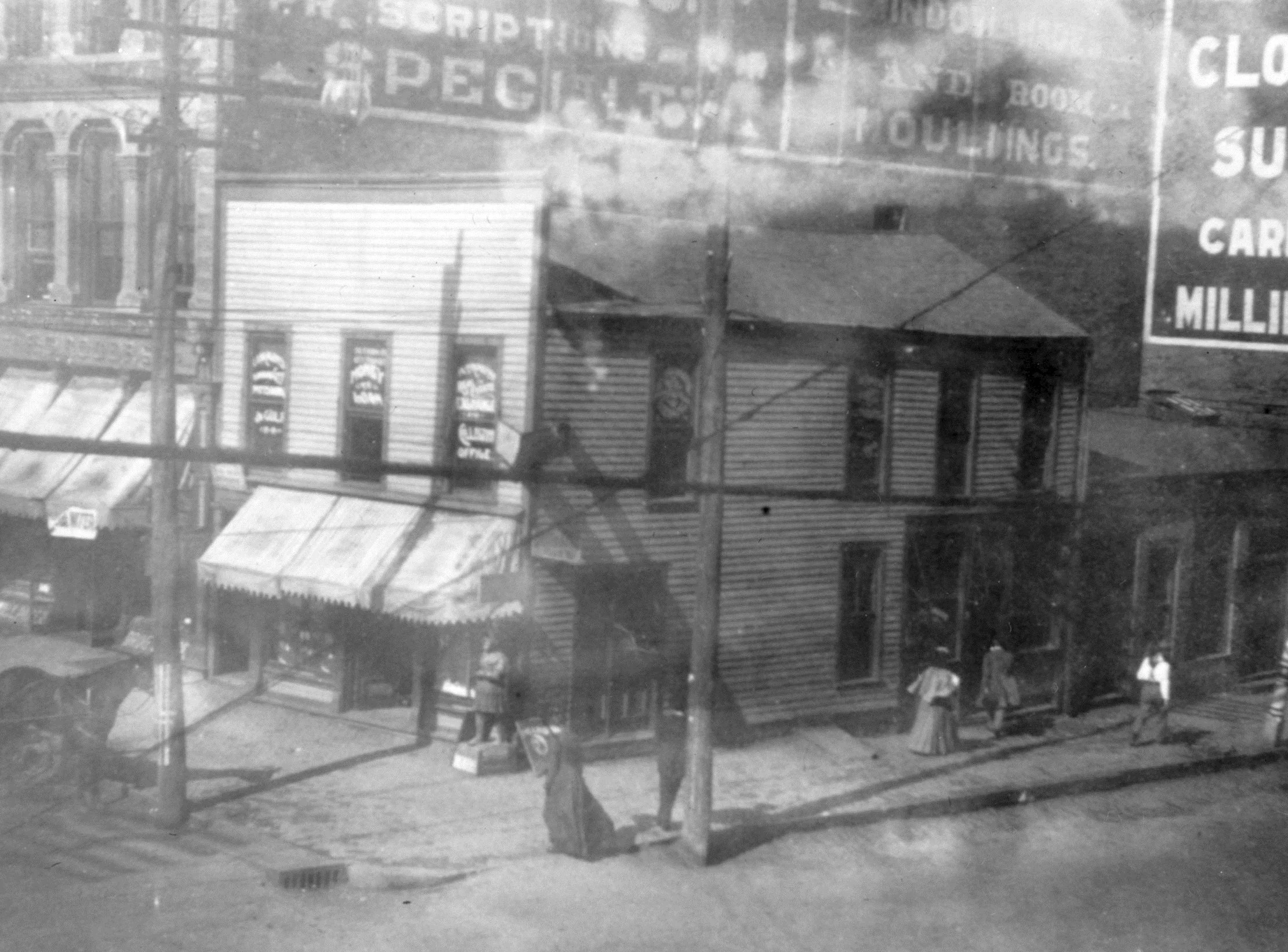 an elevated view of a street corner and building. Women in dresses walk down the sidewalk, a horse and buggy are parked on the street. Utility poles are in the foreground. Two larger brick buildings are behind the two-story frame building, each with large advertisements painted on them. The two story frame building has light colored siding, many windows, and awnings over the first story storefront.