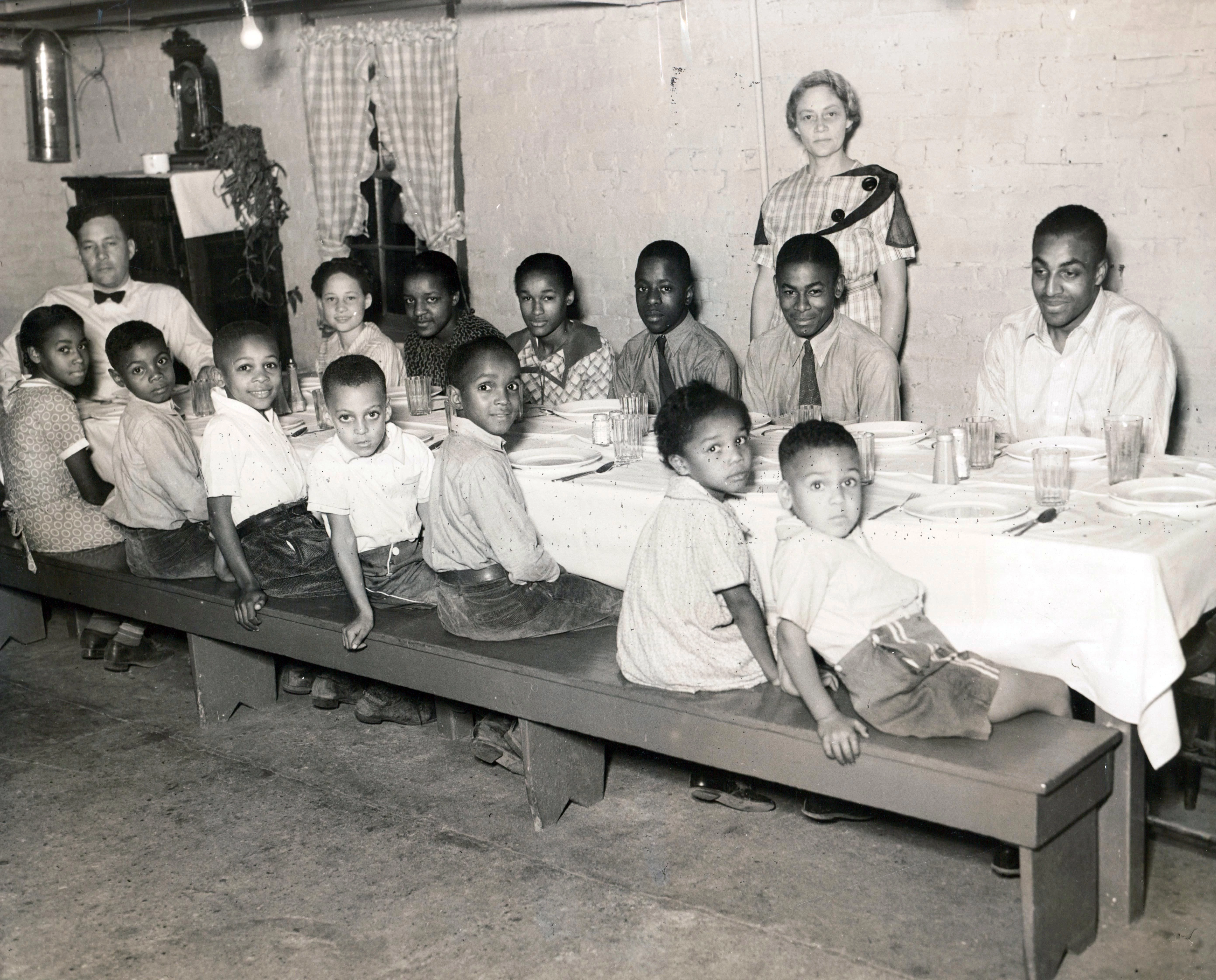 13 children and young adults sit on a long bench at a table. The table has a white table cloth and place settings for each person. One man is at the head of the table, wearing a bow tie. A woman stands behind the table in a dress. They are all looking at the camera, many of the children are smiling. All of the people in the photograph are African American. The walls appear to be lightly-painted brick.