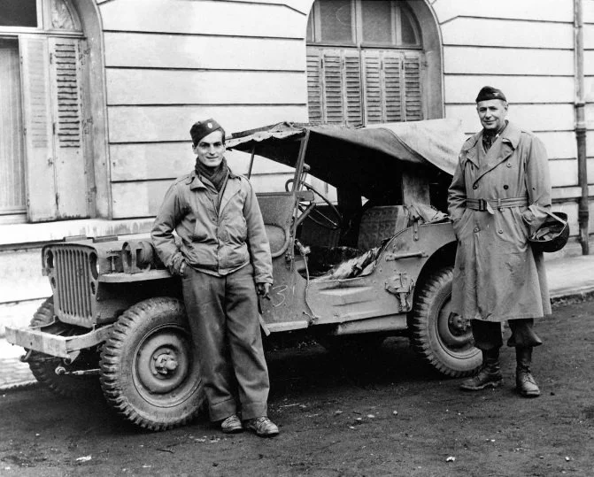 Two light-skinned men stand outside a military vehicle. They are parked on a dirt road with a masonry building with large arched windows behind them.