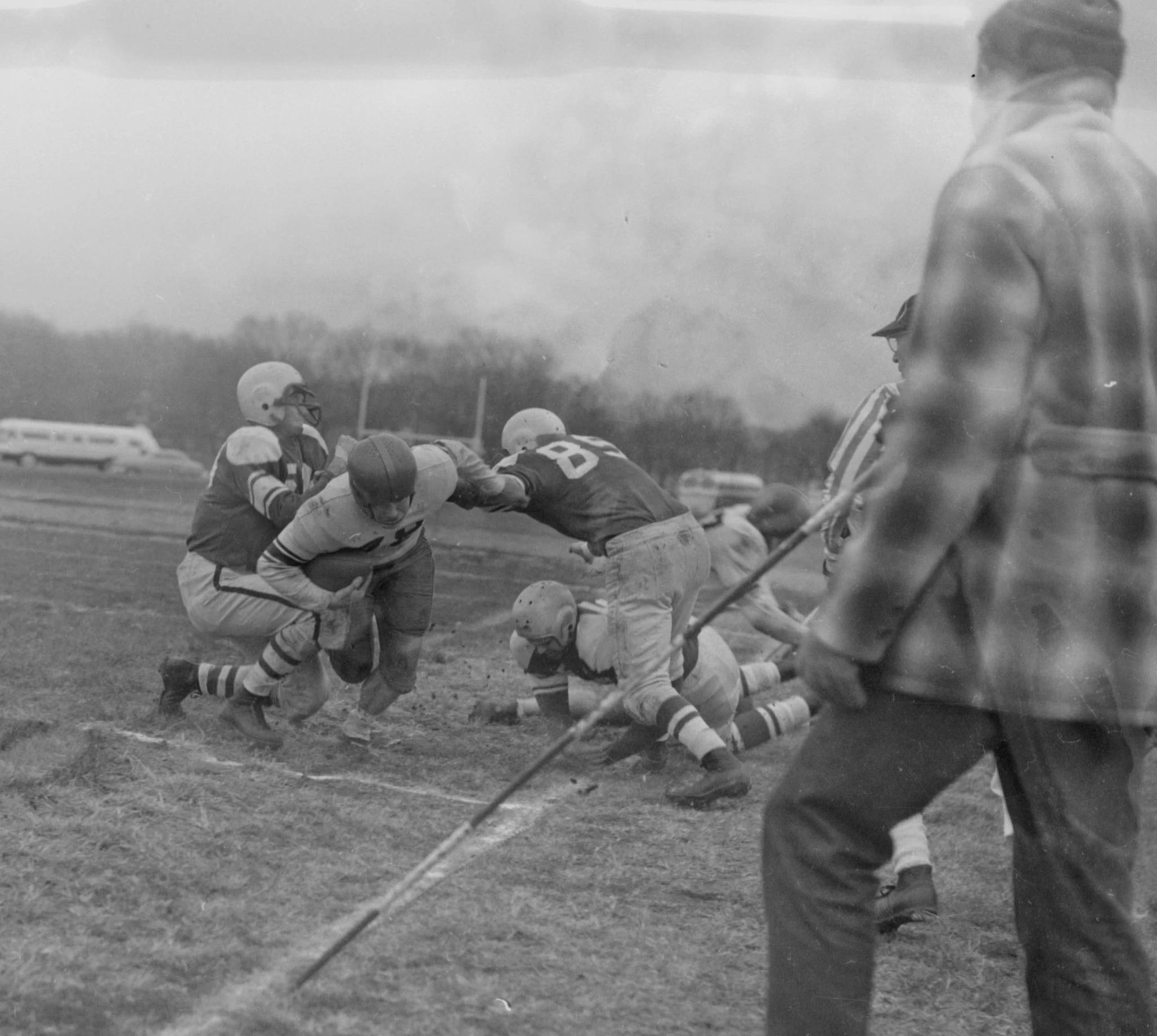 An active shot of a tackle being made on the football field. in the foreground is a man dressed in regular clothing, holding a stick.