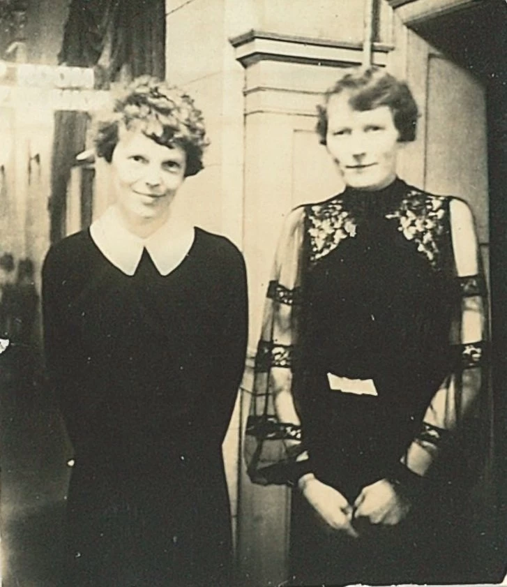 two light skinned women with short wavy hair stand next to each other looking at the camera. Amelia has a friendly closed-mouth smile and is wearing a long-sleeved black dress with large white collar. Next to her, Brenneman is wearing a dark dress with sheer sleeves.