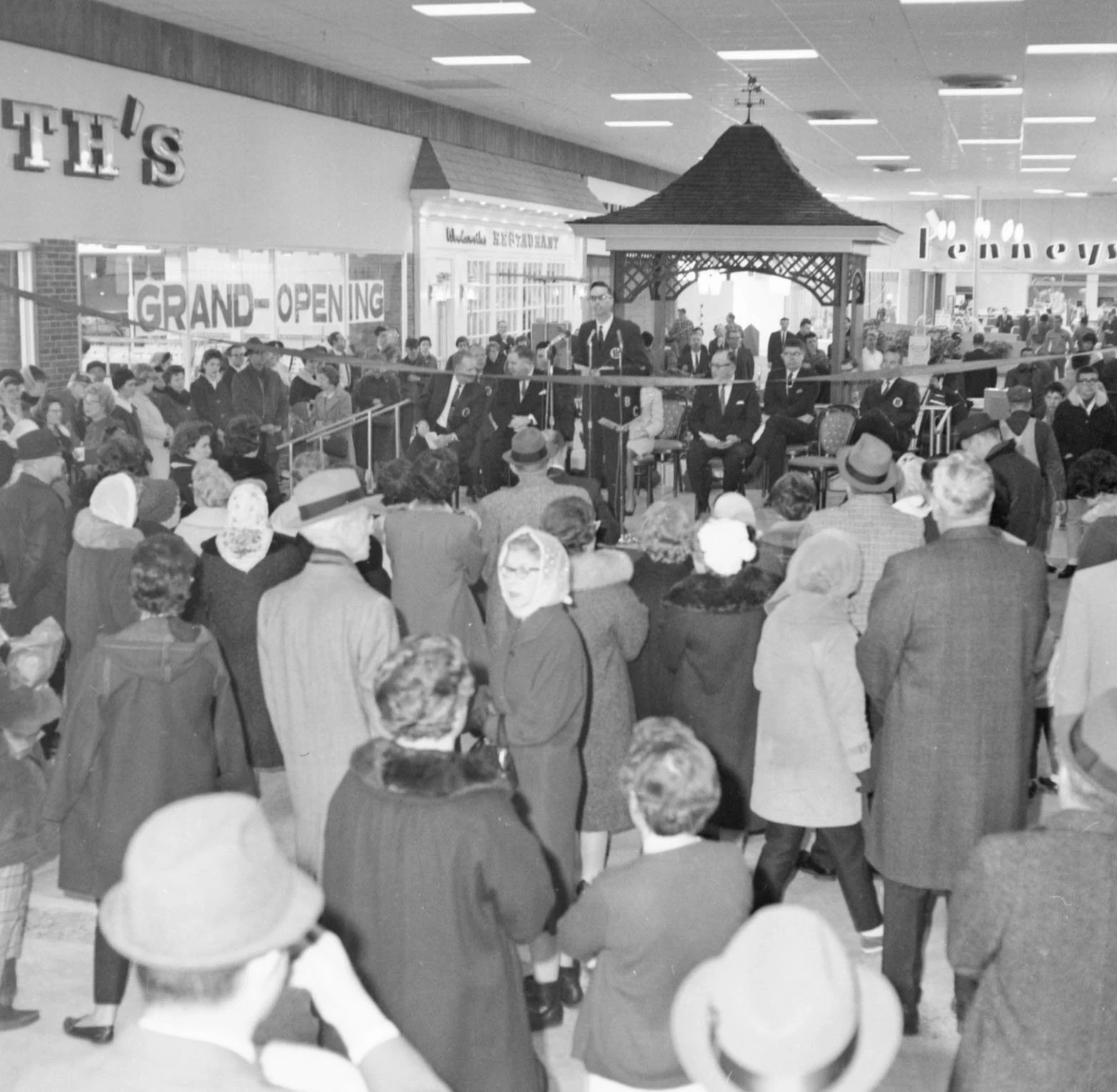 A large group of people, mostly dressed in coats and hats, are inside the mall. In the middle of the crowd is a light-skinned man wearing a suit speaking into a microphone. There is a small gazebo-like structure behind him. To the left is Woolworth's, with a sign in the window that says 