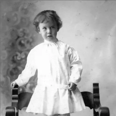 A small boy in a dress stands on a curved wood chair.