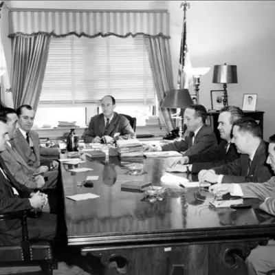 Eight men in suits sit at a table in a conference room