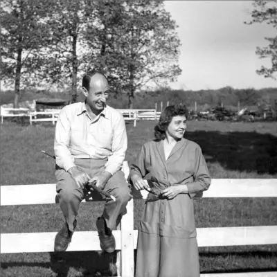 A balding man in white shirt and khaki pants sits on a fence next to a woman in a long dress.