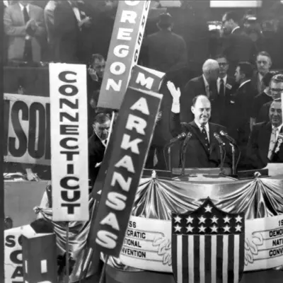 A balding man in a dark suit waves toward a crowd of men in suits. Signs are held representing various US states.
