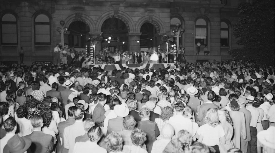 A crowd dressed in white stands in front of a stage in front of a historic courthouse