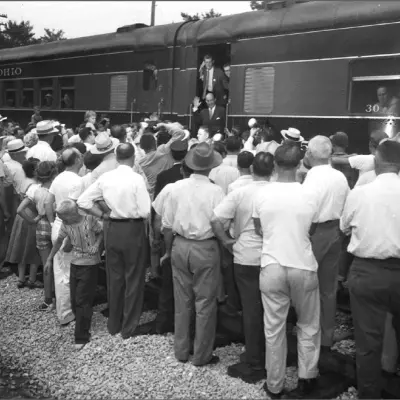 A crowd of men in white shirts, dark pants, and hats stand in front of a train. A man waves from the train.