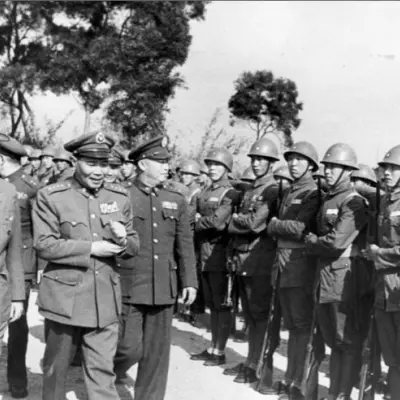 A man in a suit and a hat walks alongside Asian men in military uniforms.