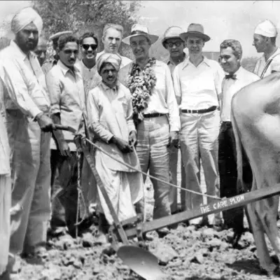 A man in a collared shirt, slacks, and a wide brimmed hat stands among other men in shirts and hats, looking at a manual plow,