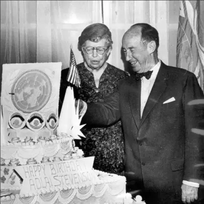 A balding man in a suit and an older woman with glasses stand next to a four tier cake decorated with flags and stars.