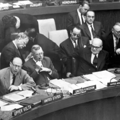 Several middle aged men in suits watch as another middle aged man in a suit speaks.