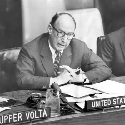 A middle aged man in glasses and a suit sits as a table and speaks into a small microphone.