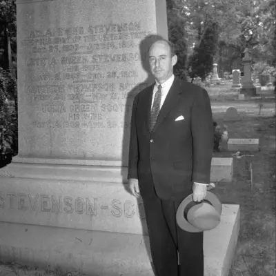 A middle aged bald man in a suit is holding a brimmed hat and standing next to a large gravestone that is taller than him.