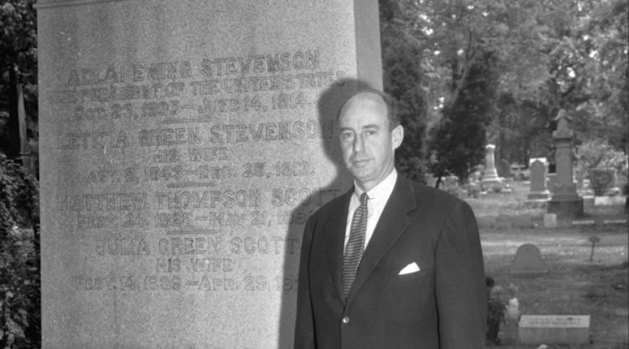 A middle aged bald man in a suit is holding a brimmed hat and standing next to a large gravestone that is taller than him.