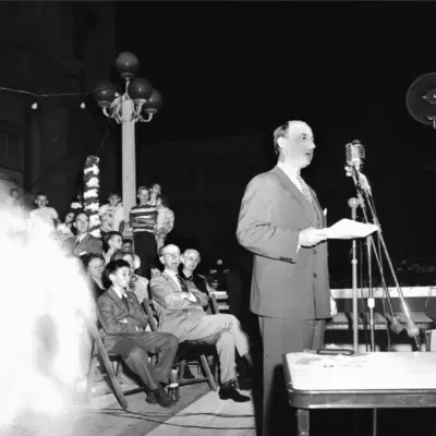 A balding man in a suit speaks into a ribbon microphone at night.