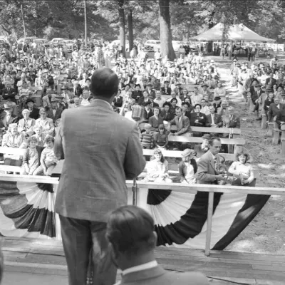 A man in a suit faces a standing crowd in a park