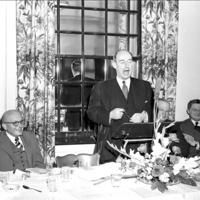 A middle aged bald man in a dark suit speaks into a microphone in front of a window.