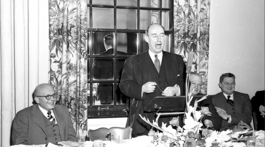 A middle aged bald man in a dark suit speaks into a microphone in front of a window.