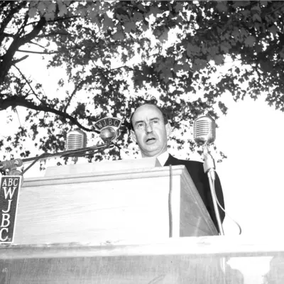 A middle aged bald man speaks into three ribbon microphones at a podium beneath trees.