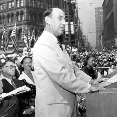 A balding man in a light colored suit stands at a podium and speaks into a microphone, surrounded by a crowd and historic buildings.