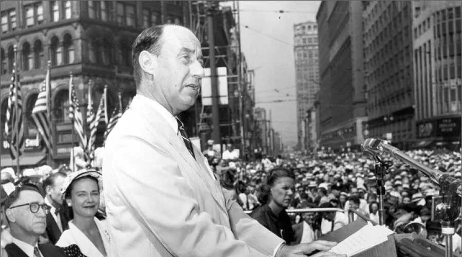 A balding man in a light colored suit stands at a podium and speaks into a microphone, surrounded by a crowd and historic buildings.