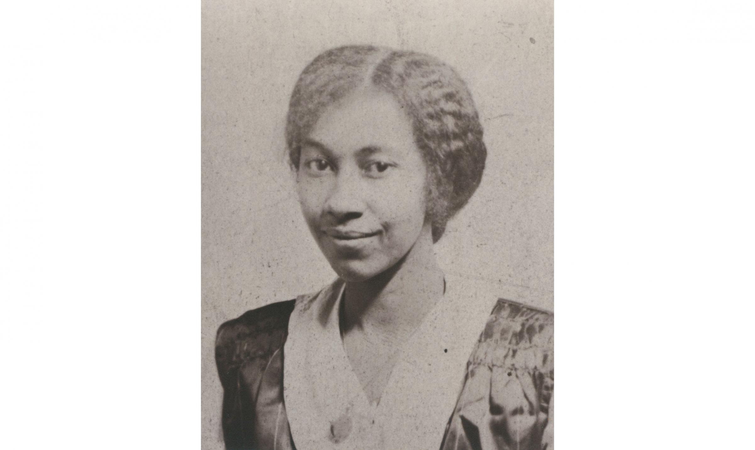Portrait of a young Black woman with her hair parted down the middle and pulled back. She is wearing a black and white dress and is looking at the camera with a closed-mouth smile.