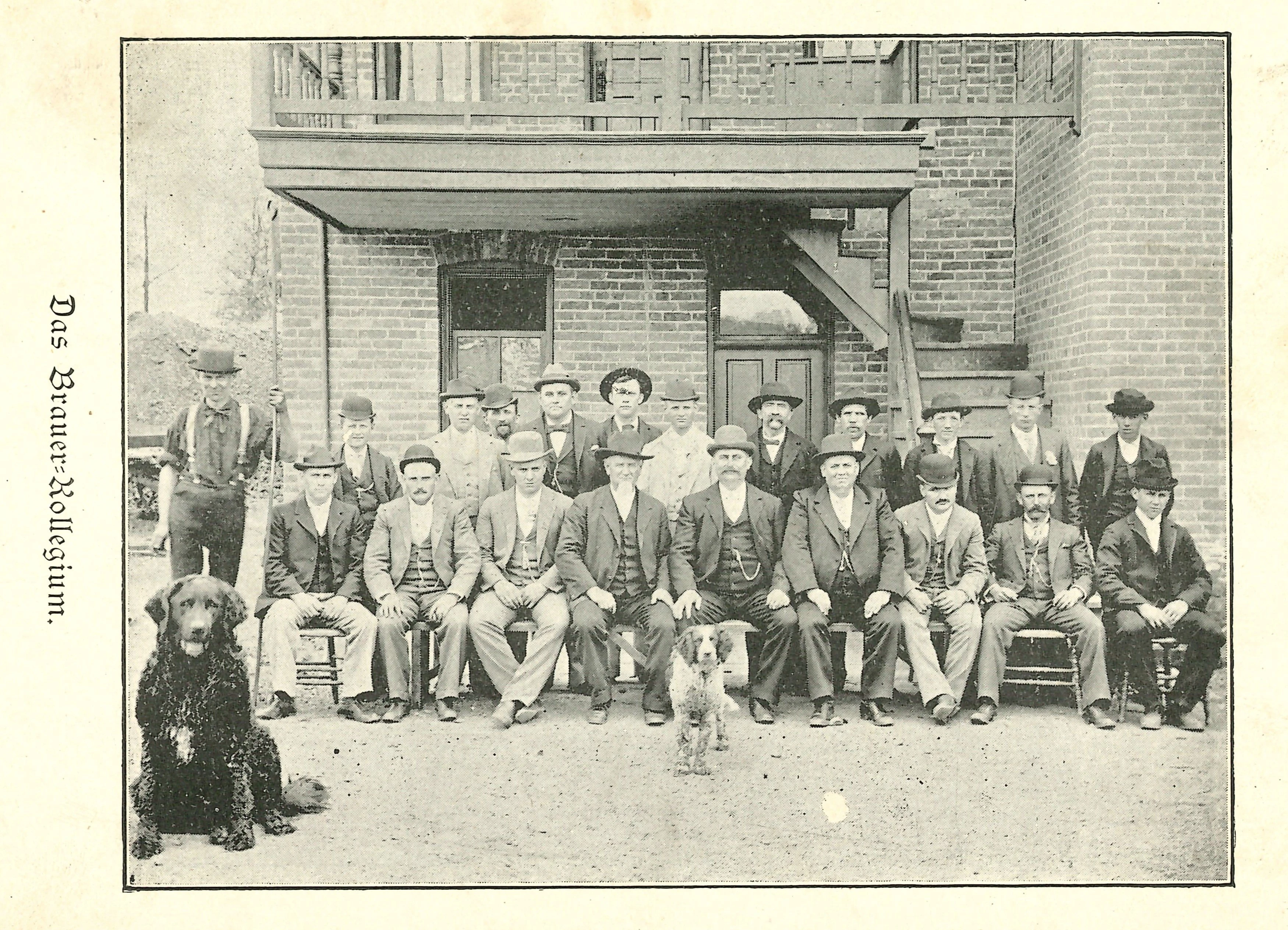 Twenty-two men sit in front of the Meyer Brewing Company accompanied by two dogs in 1893. The image appears with German text on the side that reads