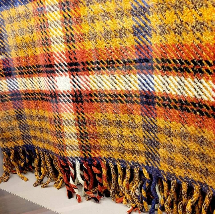 Photo of a plaid, wool blanket with fringe along the bottom. It is mostly yellow, with accents of blue, red, black, and white.