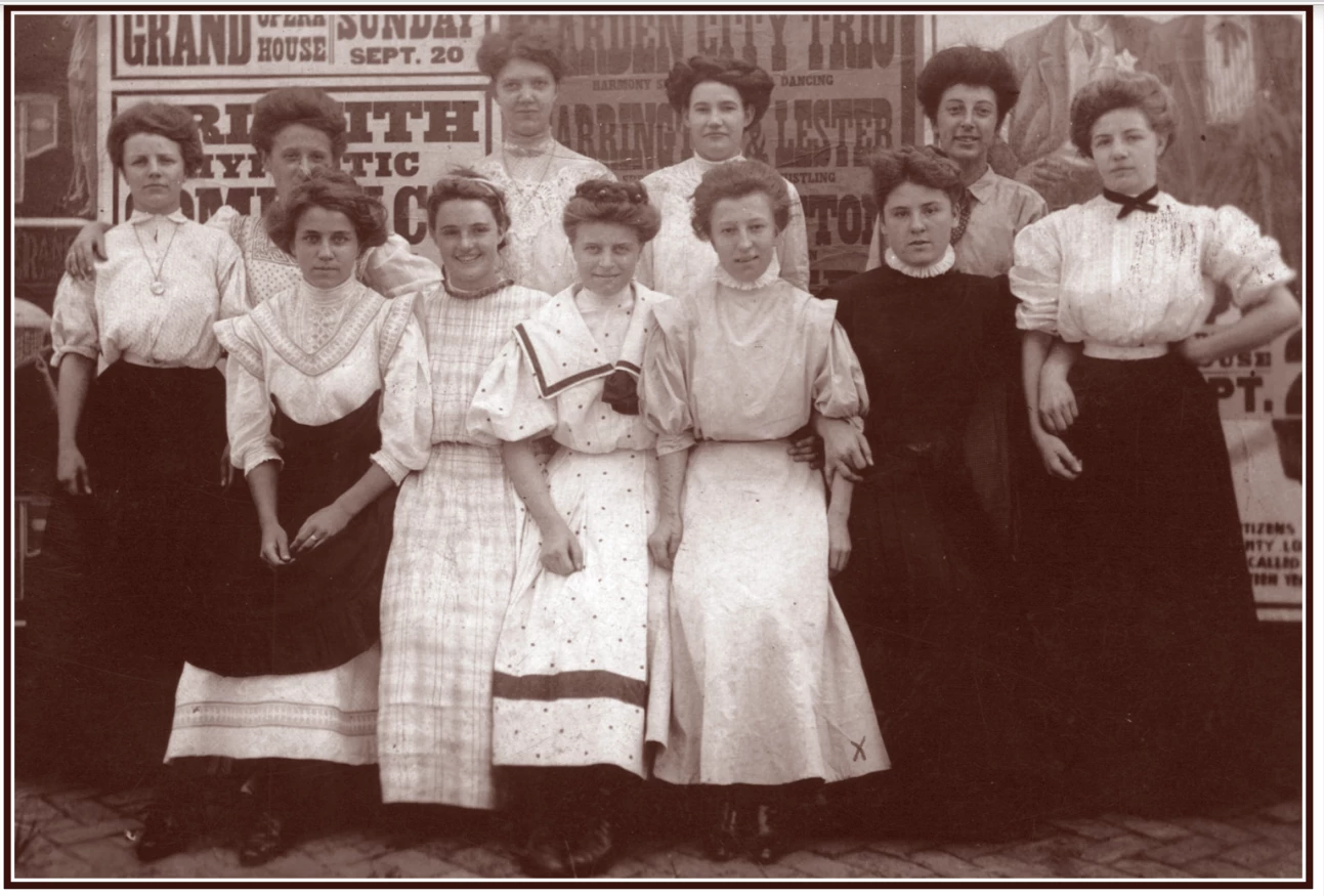 A black and white photo of 11 women standing together. They all are wearing dresses and have their hair up.