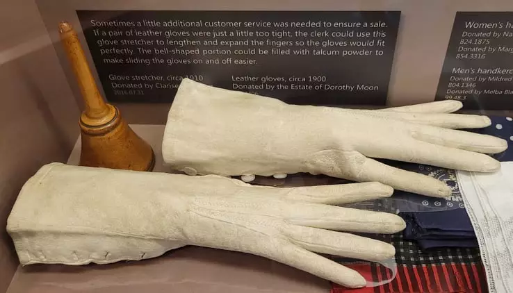 A pair of white leather gloves displayed next to a wooden glove stretching device, which consists of a flared base and shaft.