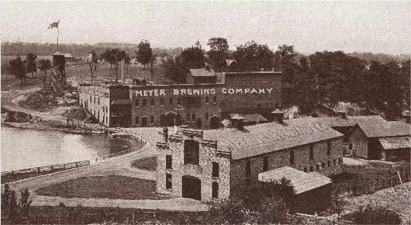 A view of a few brick buildings, one says MEYER BREWING COMPANY in big letters across the side. Trees and a water tank are in the background. On the left side of the image is a lake.