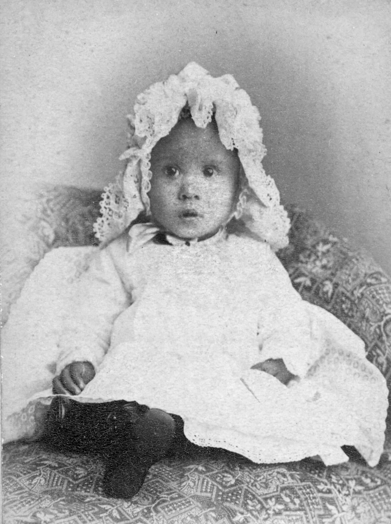 Black and white photo of an infant wearing a white frilly cap and long white dress.