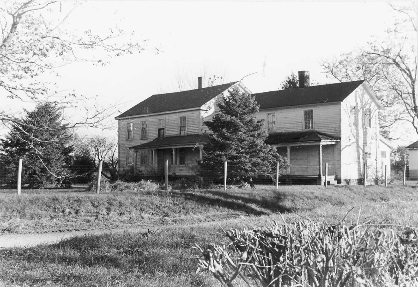 Black and white photo of a large white dilapidated farmhouse, surrounded by trees and a wire fence.