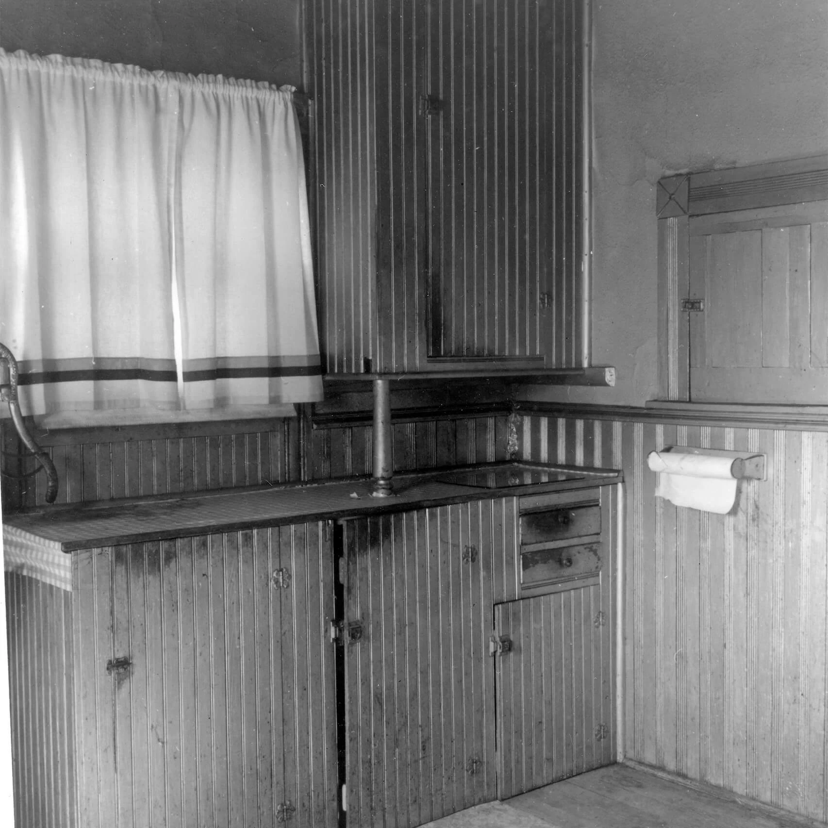 Black and white photo of a kitchen. It is almost entirely wood paneling, with a vertical pattern. There are white curtains in the upper left corner and the handle of a well pump is just visible on the left edge. There are several cabinet doors and a paper towel holder.