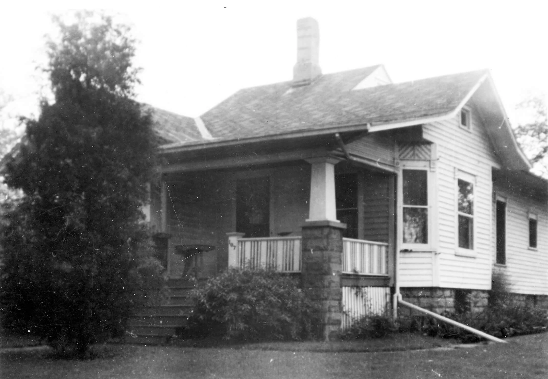 Black and white image of a single-story light colored frame house with small front porch and chimney.
