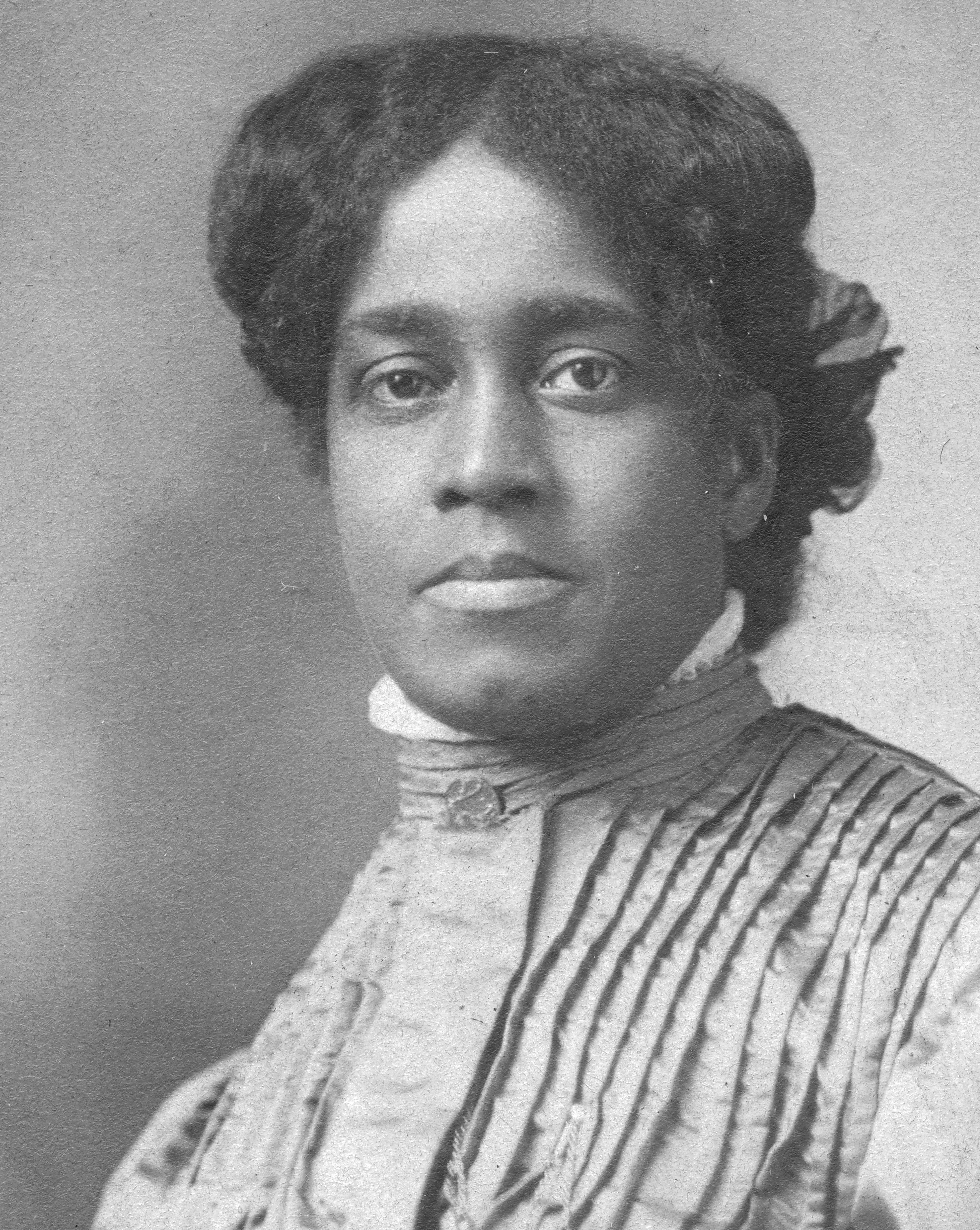 Black and white photo of a Black woman looking confidently into the camera. Her hair is parted down the middle and pulled back. Her dress comes up to her neck and is silk or another kind of shiny fabric, with many ironed pleats.
