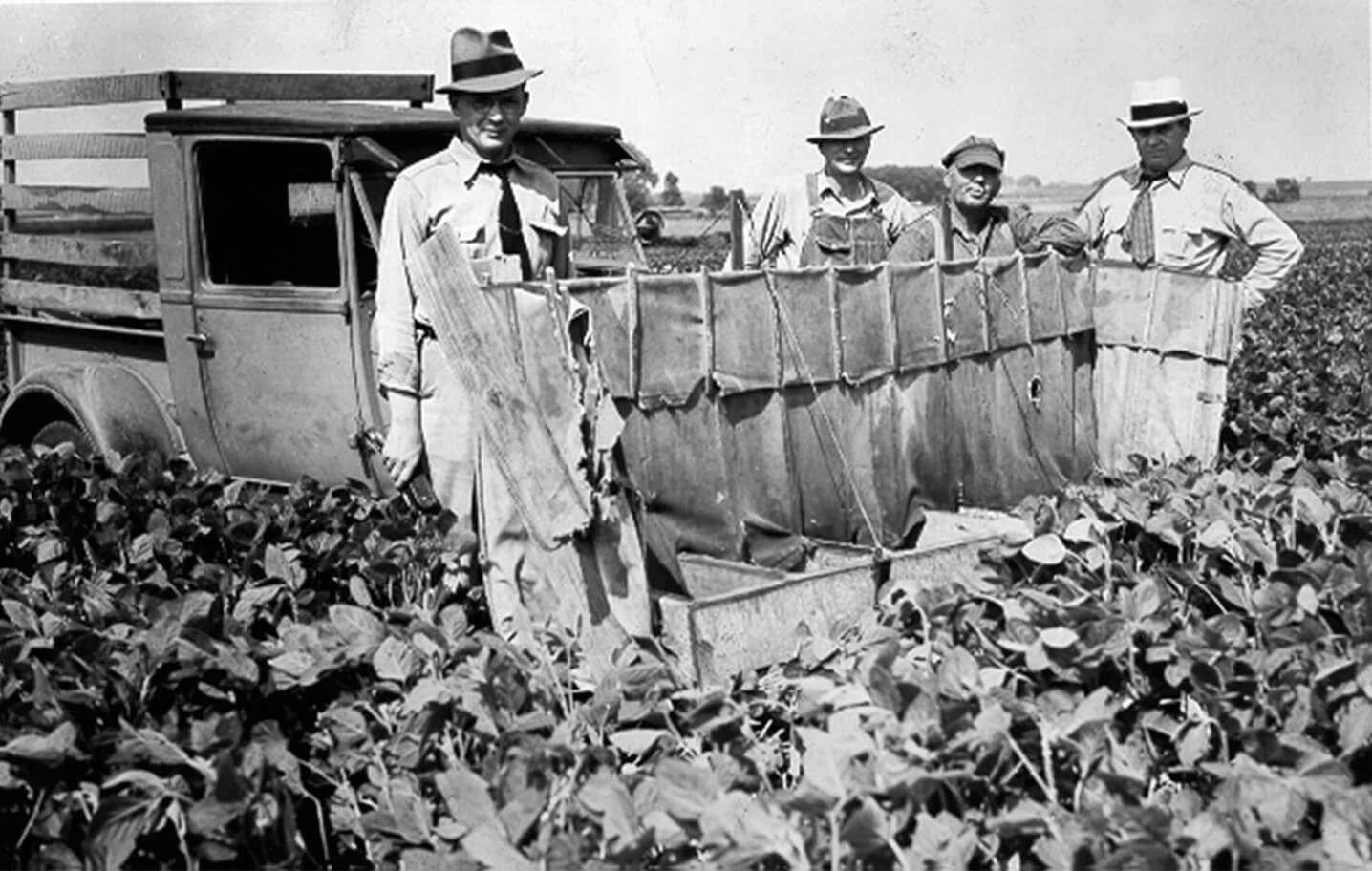 Four men in a variety of hats, collared shirts, and ties stand in front of a 1940s truck in a field. The truck has a metal frame welded to the front and a fabric curtain.