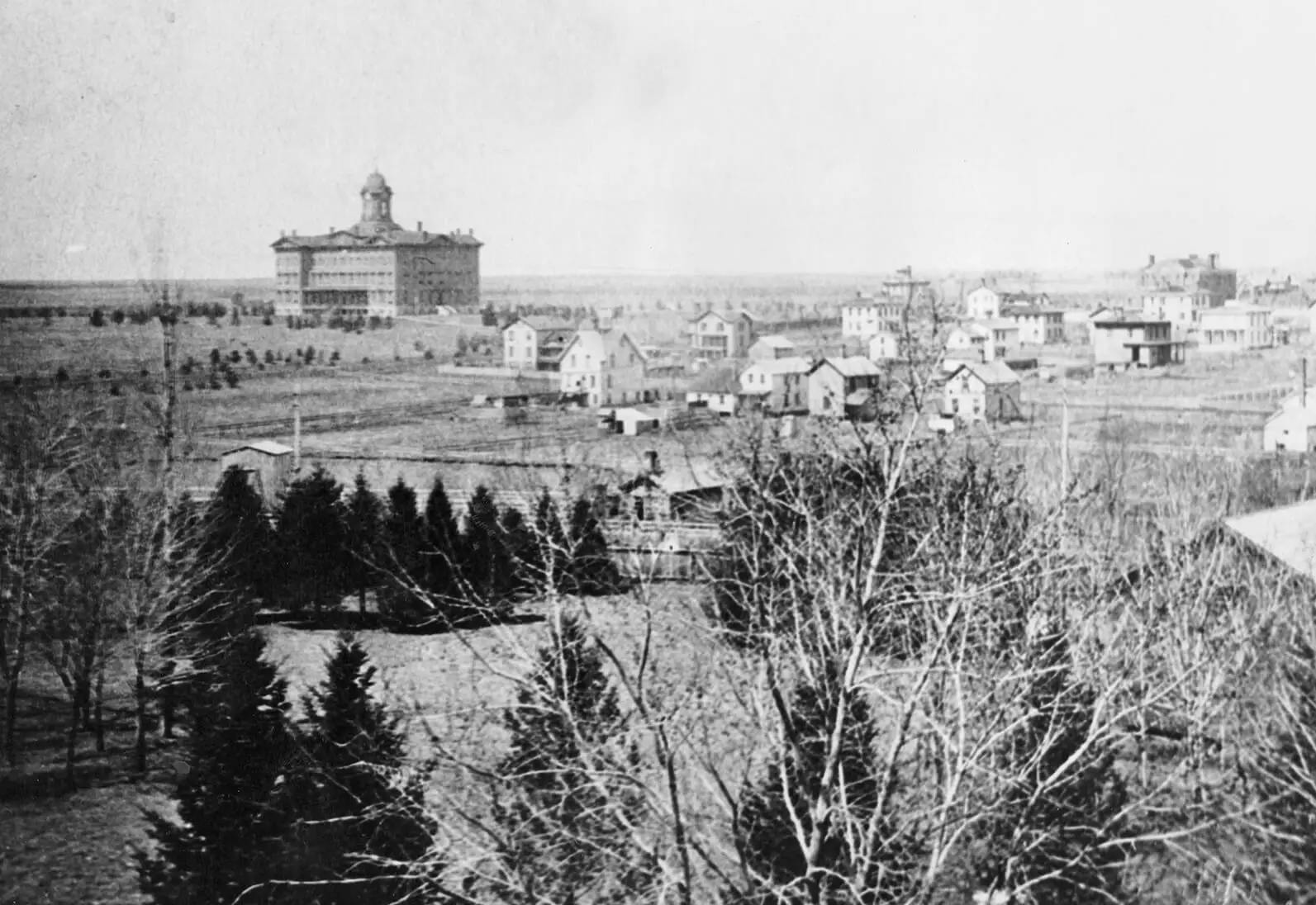 This black and white photograph shows Black and white photo of a large multi-story building in the distance and smaller structures in the surrounding area. Many trees are in the foreground.