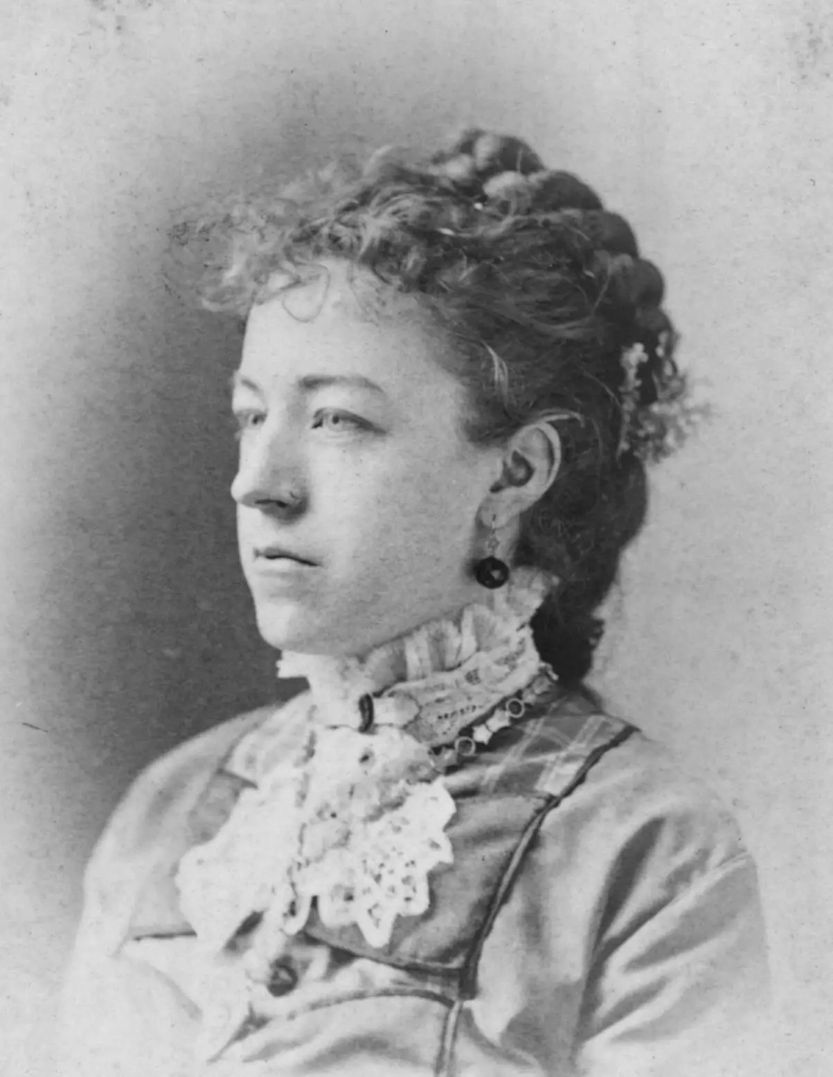 Portrait of Alice Fell, a white woman wearing a long-sleeve dress with lace collar, charm necklace, and gem-like earrings. Her dark hair is pulled back and she has a serious look.