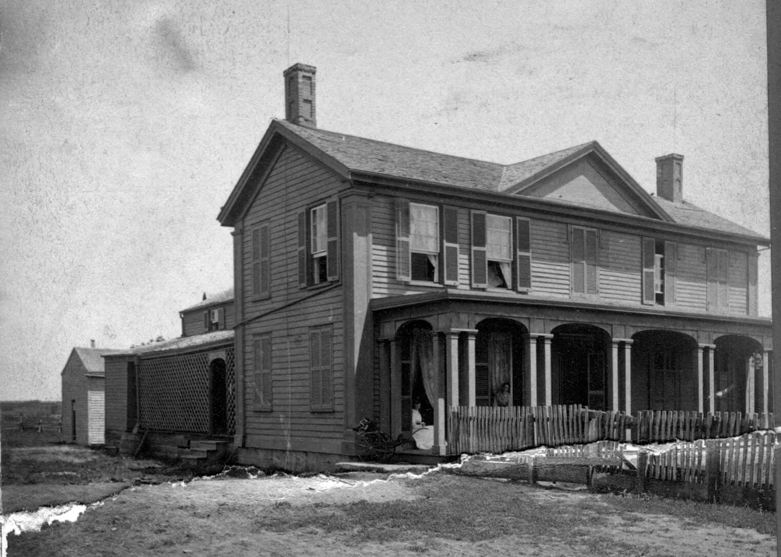 Black and white photo of a two-story house with a front porch and picket fence. Some upper windows are open, and a young woman in a white dress sits in the doorway.