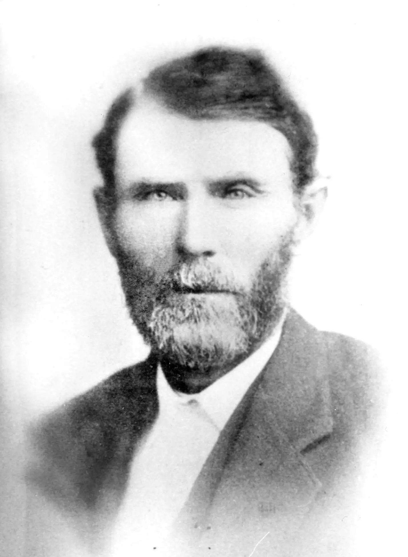 Black and white photograph of a white man with a salt and pepper beard and dark hair combed to the side. he is wearing a suit and white collared shirt.