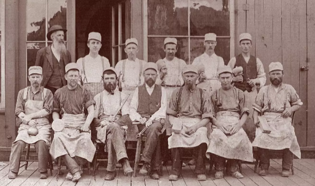 Portrait of a group of 13 men, dressed in work shirts, suspenders, soft caps, and aprons. Some men are holding small pots. One of the men is dressed in a suit and rounded hat. The first row is seated and the second row is standing behind them. They are standing in front of a building.