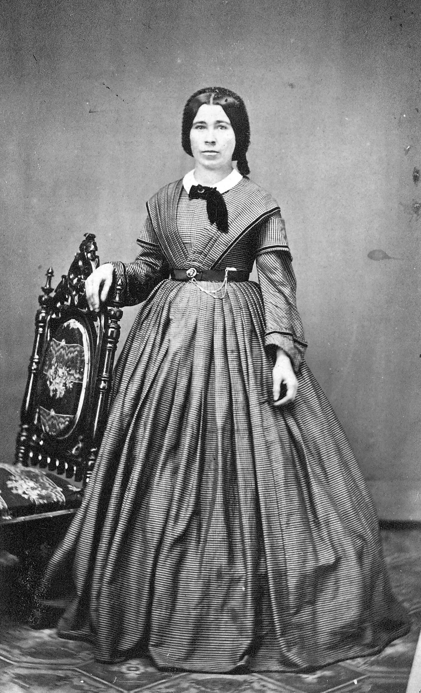 Full-length black and white portrait of a white woman standing in a floor-length dress. The dress is horizontally pin-striped. Her dark hair is parted down the middle and her arm is resting on a tall chair.