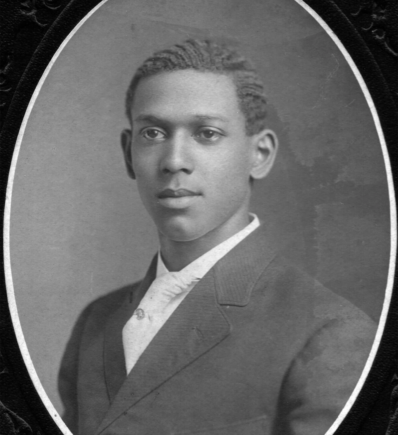 Black and white portrait of a young Black man with short hair, large ears, and wearing a dark striped suit and white shirt.