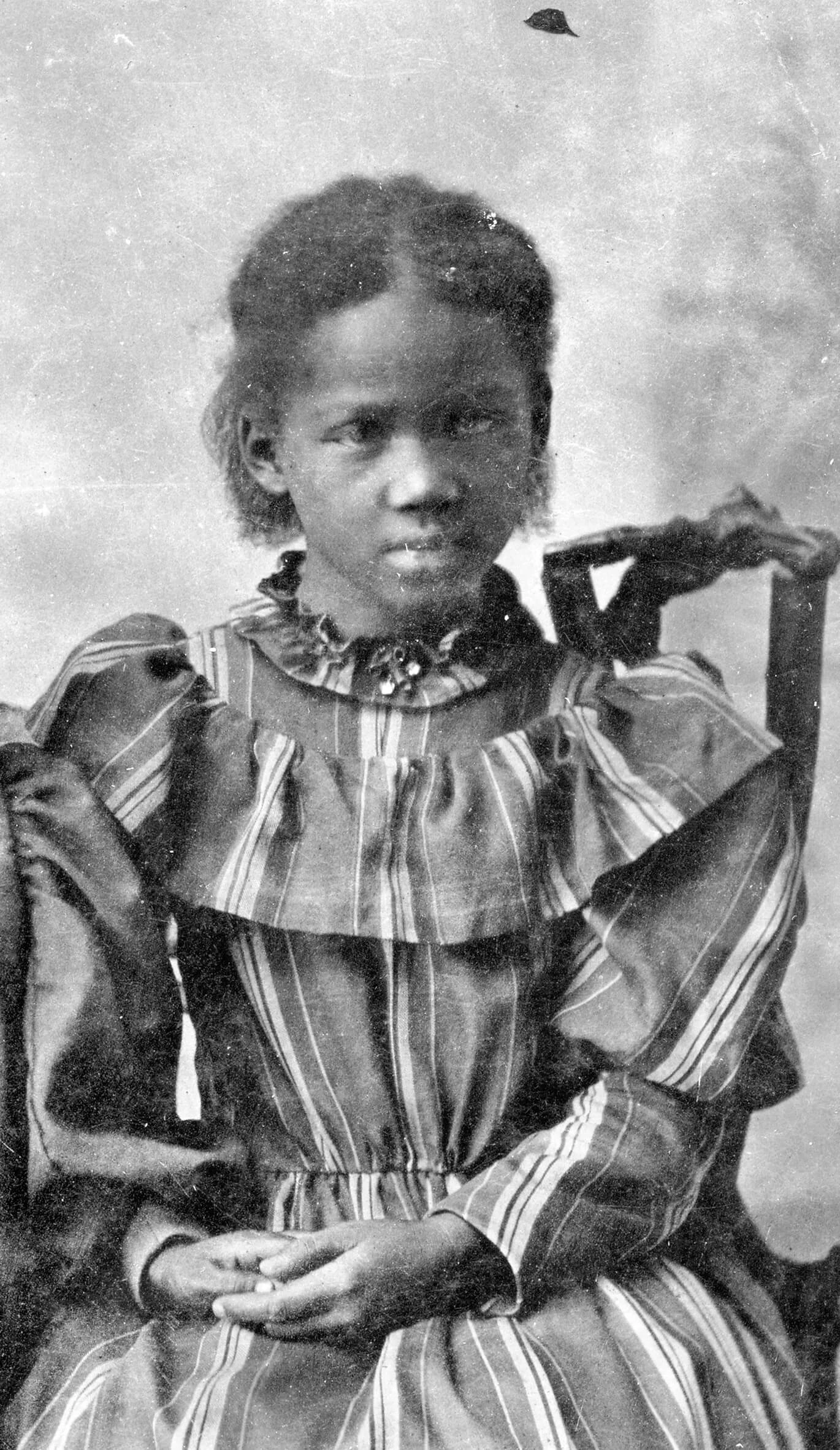 Portrait of a young Black girl wearing a striped dress. Her hands are together on her lap