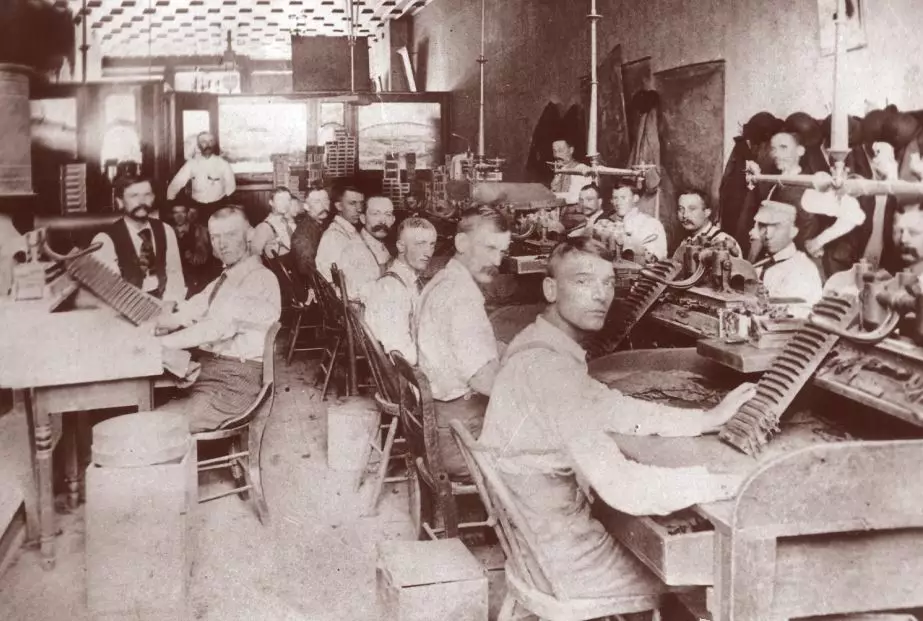 Portrait of several men at work in a cigar factory. They are seated at wood desks with drawers, facing each other. All of them appear to be light-skinned and about half have handlebar mustaches. Nearly all of the men are wearing overalls and light colored shirts. Two men are wearing suits.
