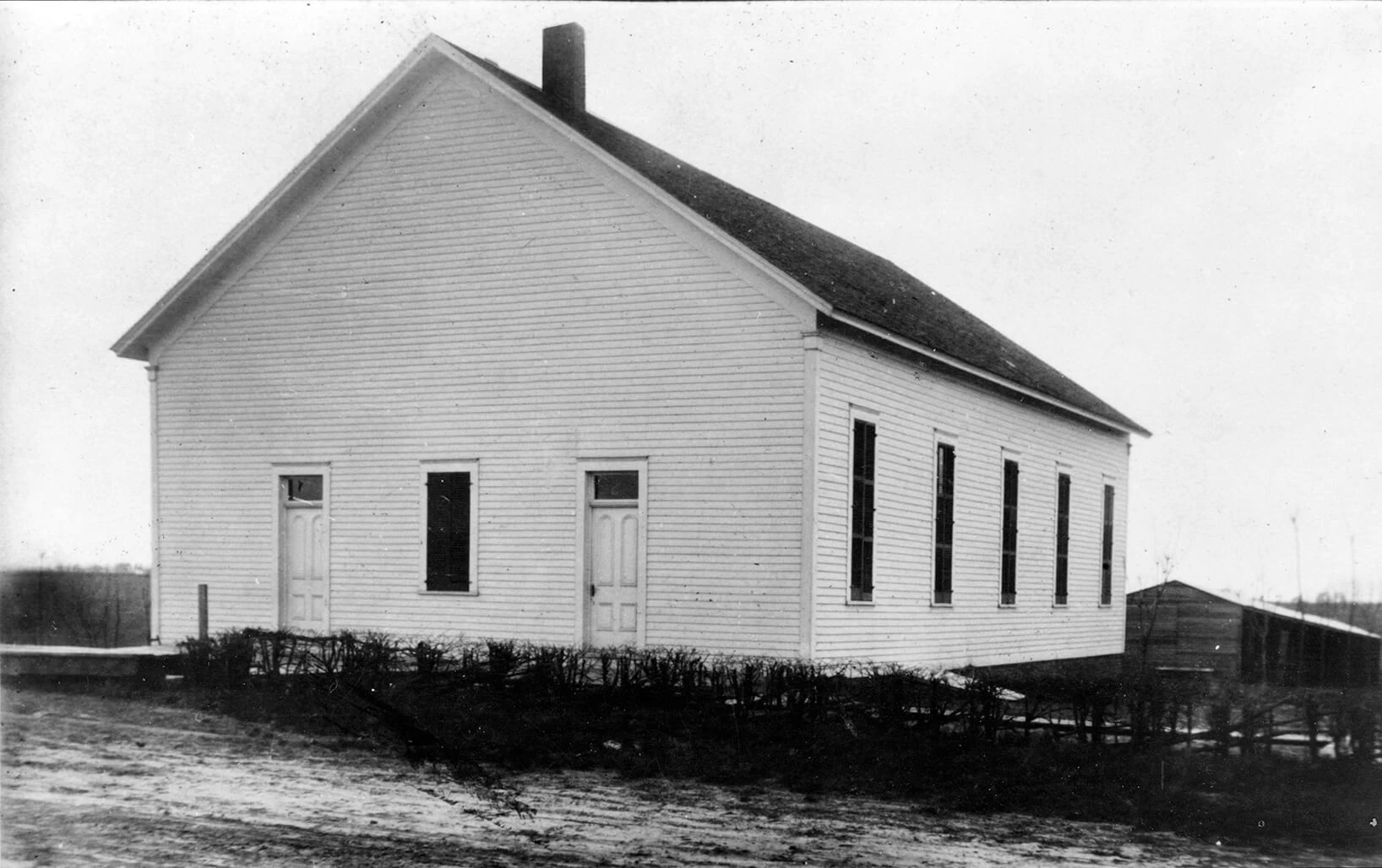 Black and white photo of a wooden church with a high pitched roof and chimney. Two doors and a window on the front, five windows on the side. Low shrubbery in the foreground.