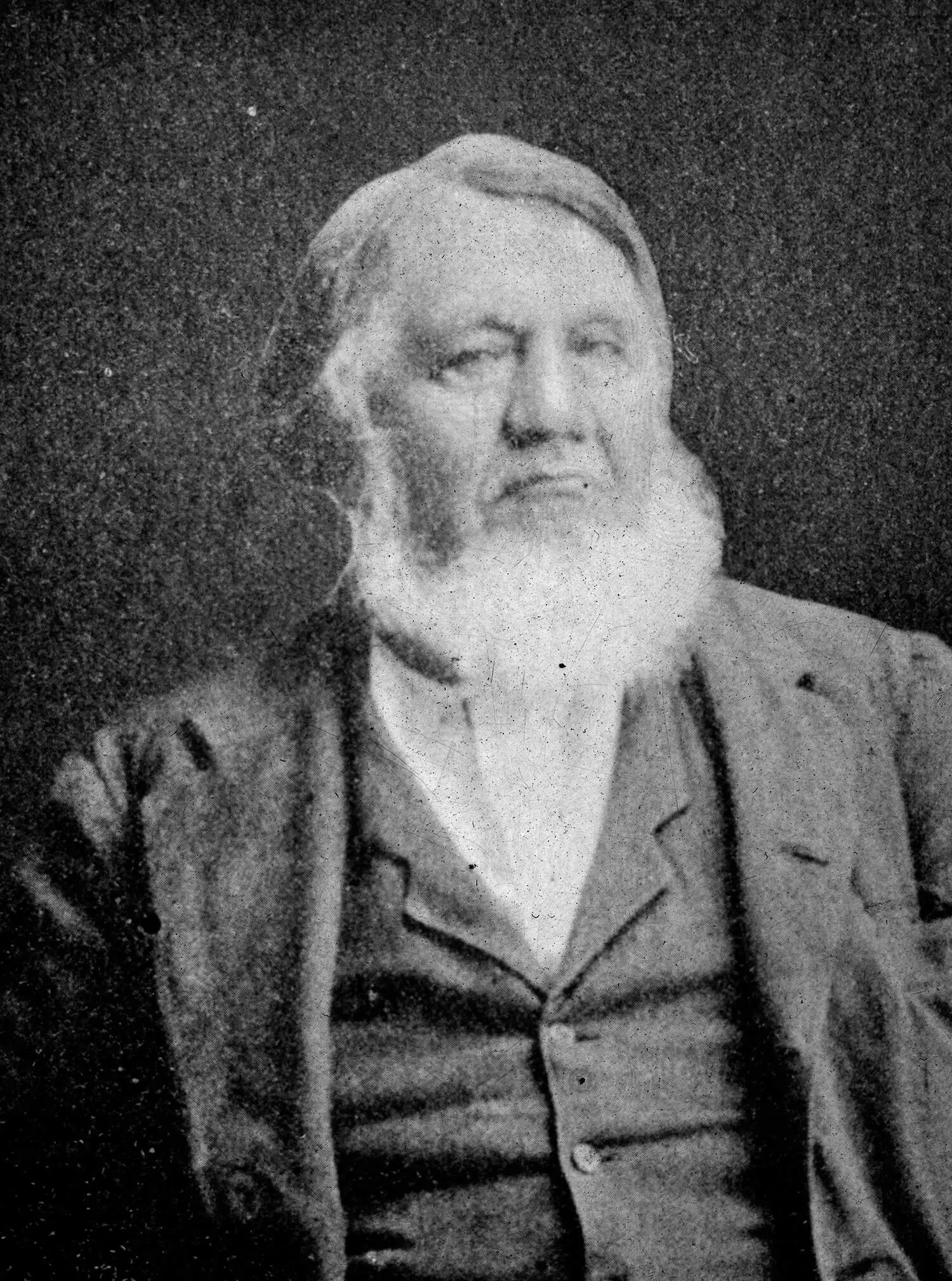 A grainy black and white photo of a white man with a white beard and white hair. He is wearing a white shirt, dark vest, and jacket.